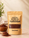 A stand-up pouch of Confectoland Cocoa Powder in a kraft paper style package with a clear window showing the dark brown cocoa powder inside. The pouch is photographed on a light beige surface with cocoa powder spilled in front, a wooden bowl, and a small plant in the background.