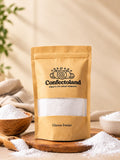 A kraft paper stand-up pouch of Confectoland Glucose Powder stands on a wooden board with scattered glucose powder around it. The pouch has a clear window showing the glucose powder inside and black printed branding and text.