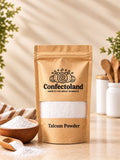 A kraft paper stand-up pouch labeled Confectoland Talcum Powder stands on a tabletop with a small wooden bowl and scoop of talcum powder beside it. The background is softly blurred with plants and kitchen utensils, creating a warm, styled setting.