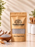A kraft paper stand-up pouch of Confectoland Silver Sugar Pearls stands on a light countertop with a window-box style kitchen scene. The pouch has a clear window showing the silver sugar pearls and the brand name 'Confectoland' printed on the front.