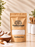A kraft paper stand-up pouch of Confectoland Xanthan Gum stands on a light countertop with a bowl of Xanthan Gum powder and a wooden spoon nearby. The background is softly blurred with plants and kitchen utensils, creating a warm, natural setting.